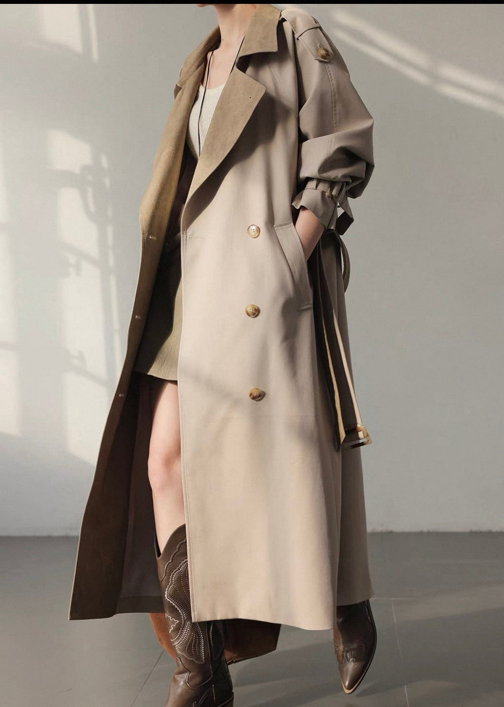 A model wearing a long beige trench coat over a white dress, styled with brown leather boots, standing in a minimalist studio with soft window shadows.