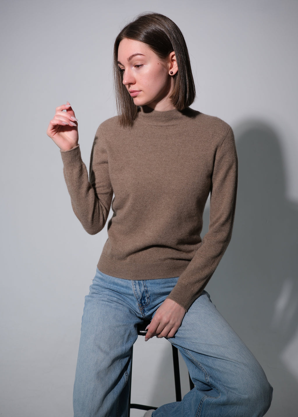 A woman wearing a brown mock-neck knit sweater and light blue jeans, sitting on a stool against a plain gray studio background.