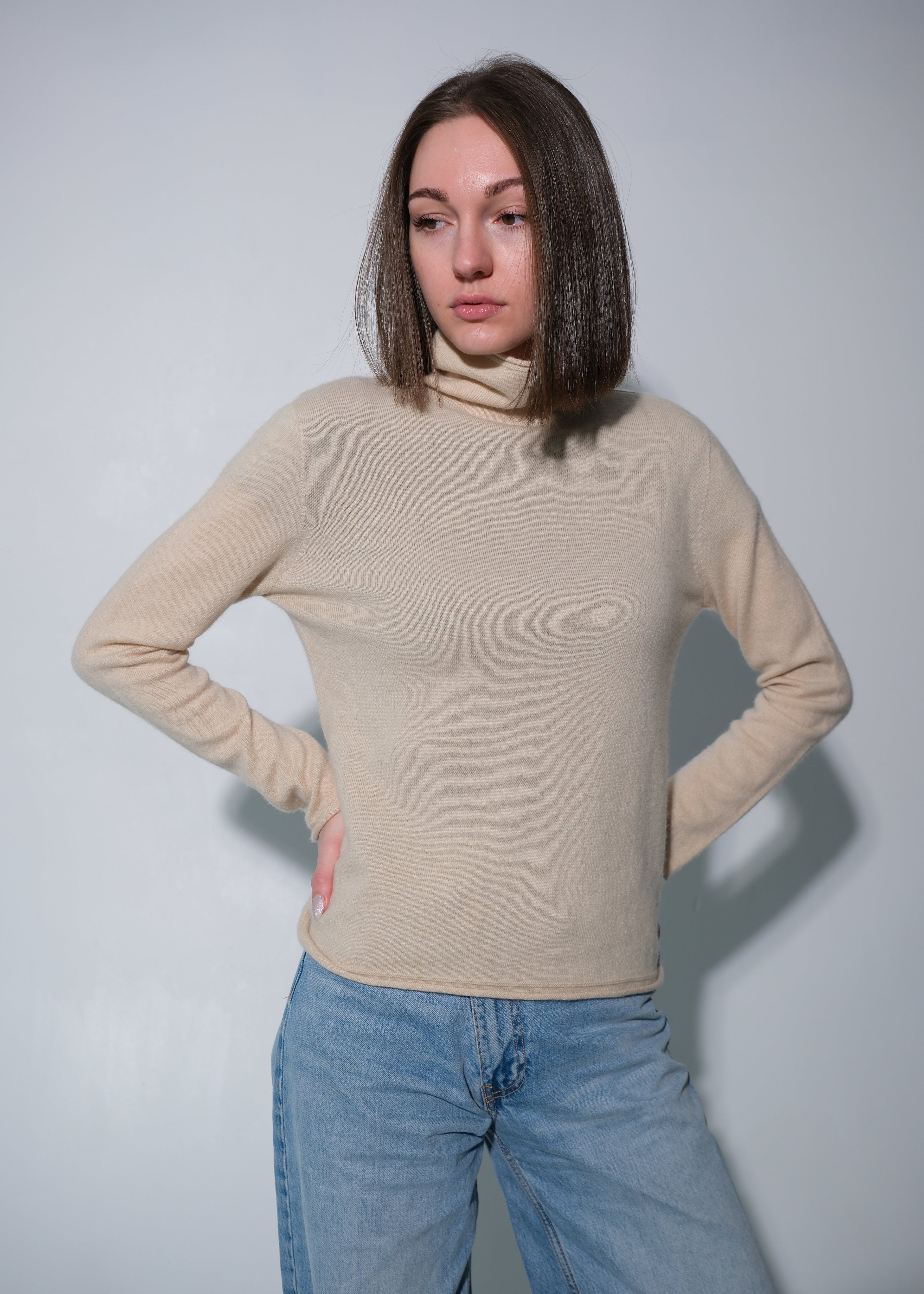A woman with shoulder-length brown hair wearing a beige turtleneck sweater and light blue jeans, posing against a light gray studio background.