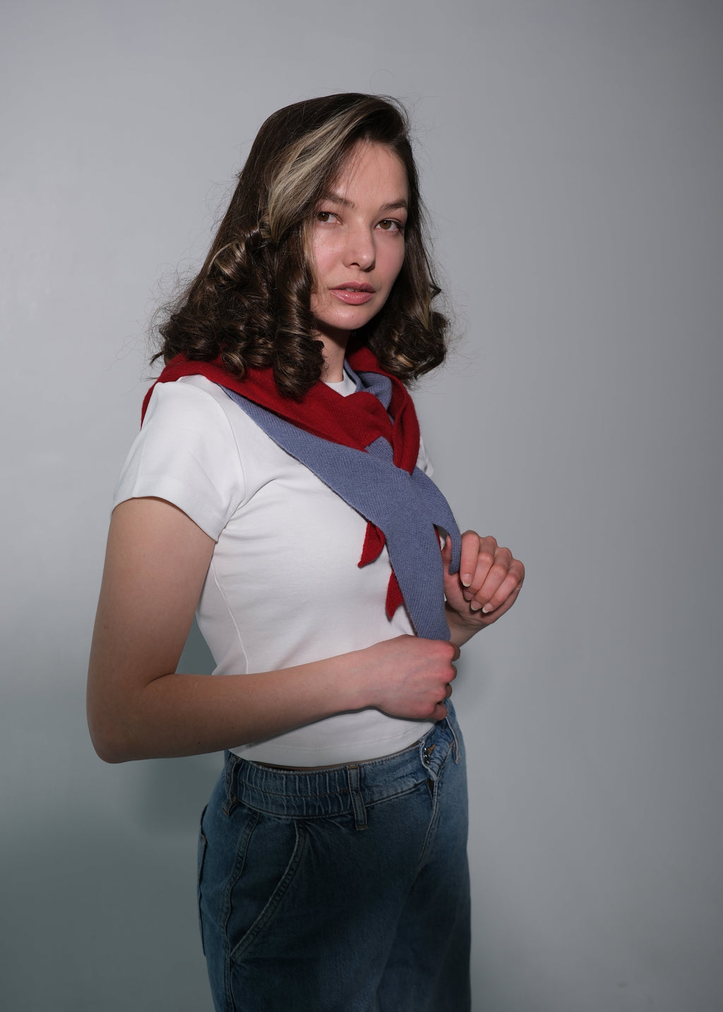 A woman modeling a unique gray shoulder strap scarf with red accents and a white t-shirt, paired with blue jeans, against a plain gray studio background.