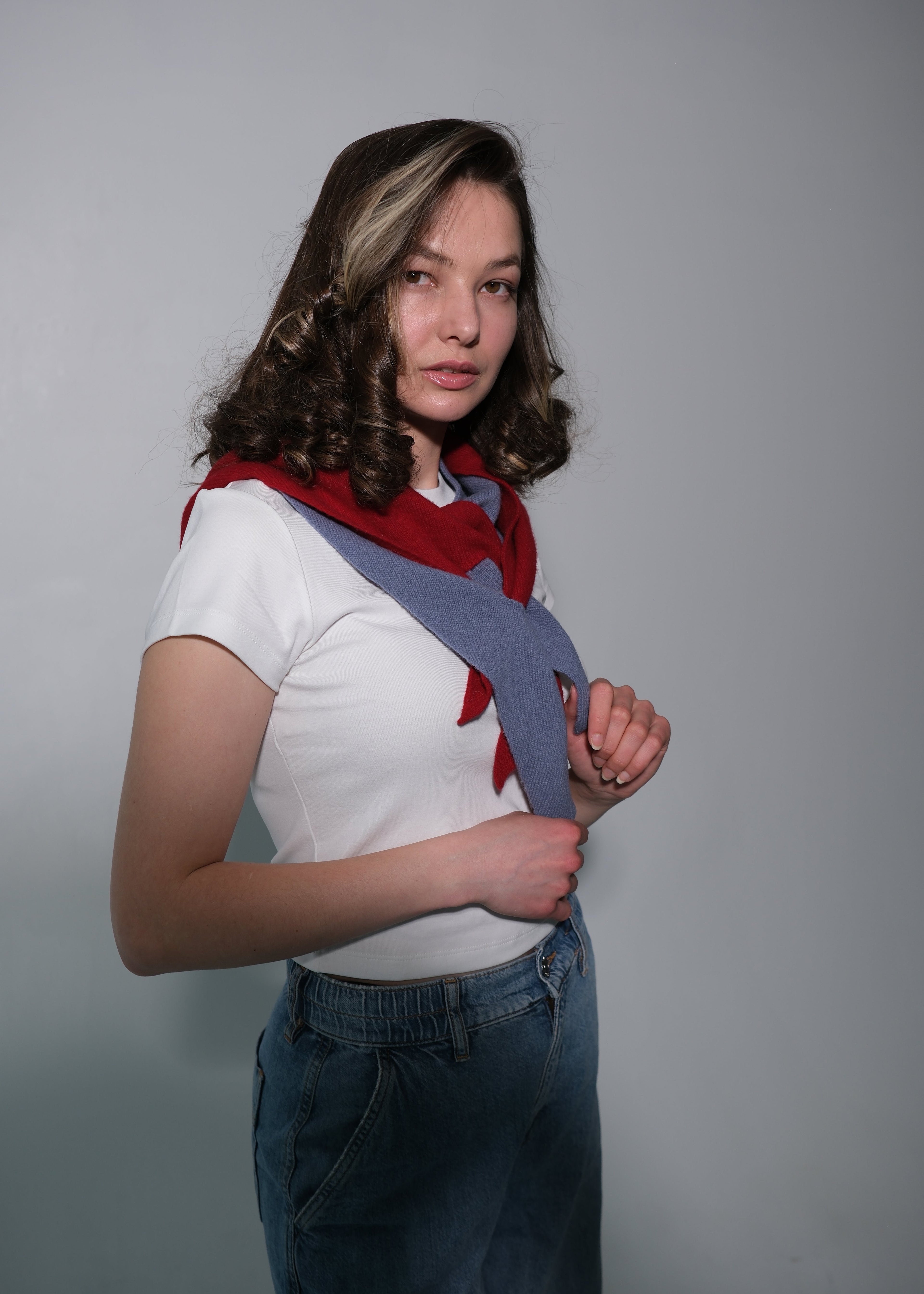 A woman modeling a unique gray shoulder strap scarf with red accents and a white t-shirt, paired with blue jeans, against a plain gray studio background.