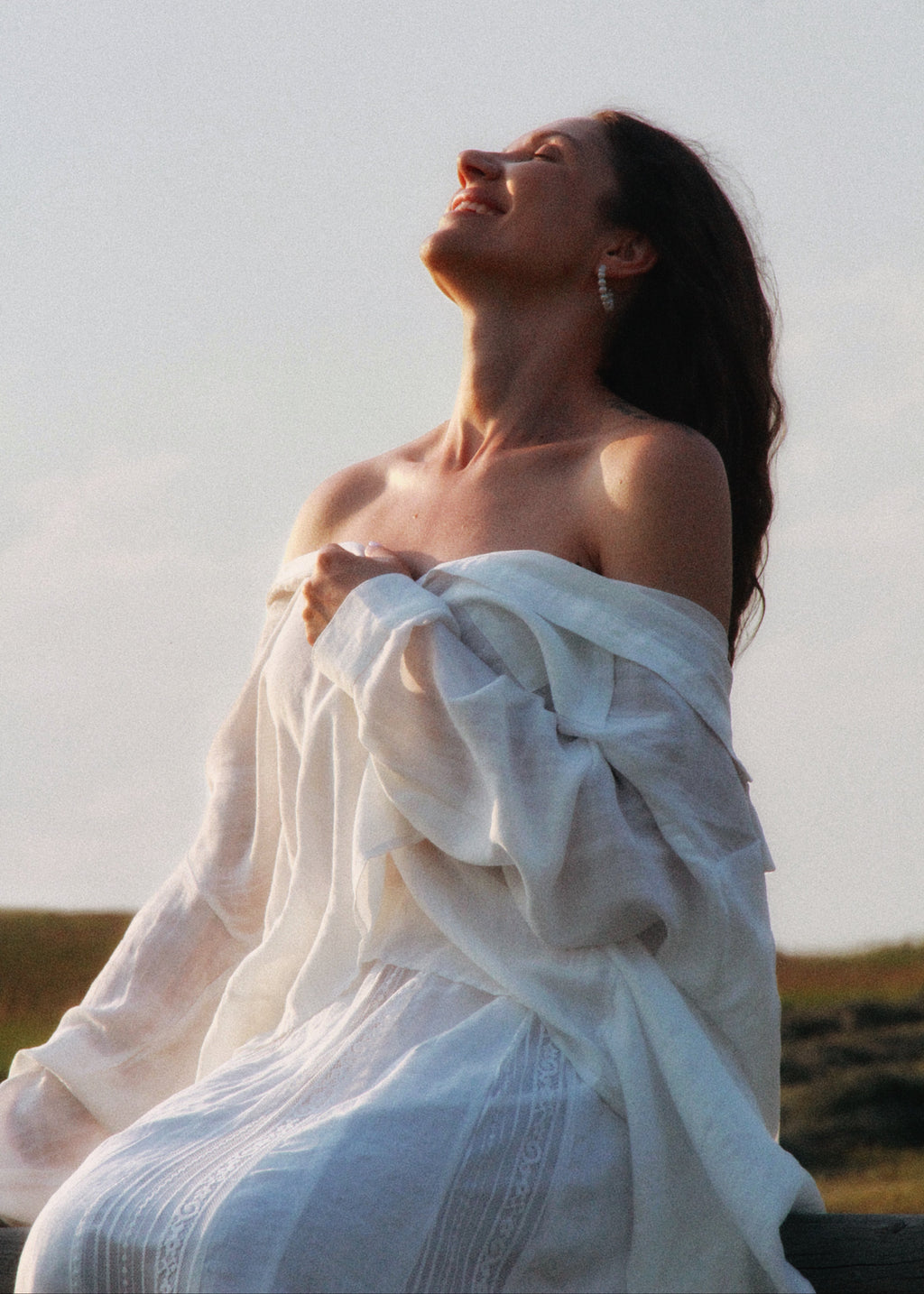 A woman outdoors wearing a flowing white off-the-shoulder dress or robe, with her head tilted back in a warm natural light. The background is a soft-focus pale sky and low landscape.