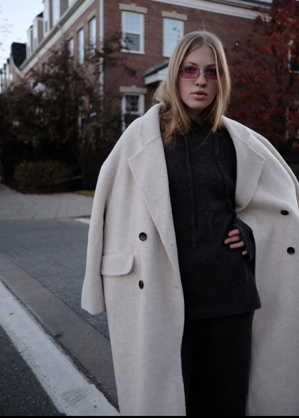 A woman standing on a street wearing a long cream-colored double-breasted coat over a black outfit, with pink-tinted sunglasses and a brick house in the background.